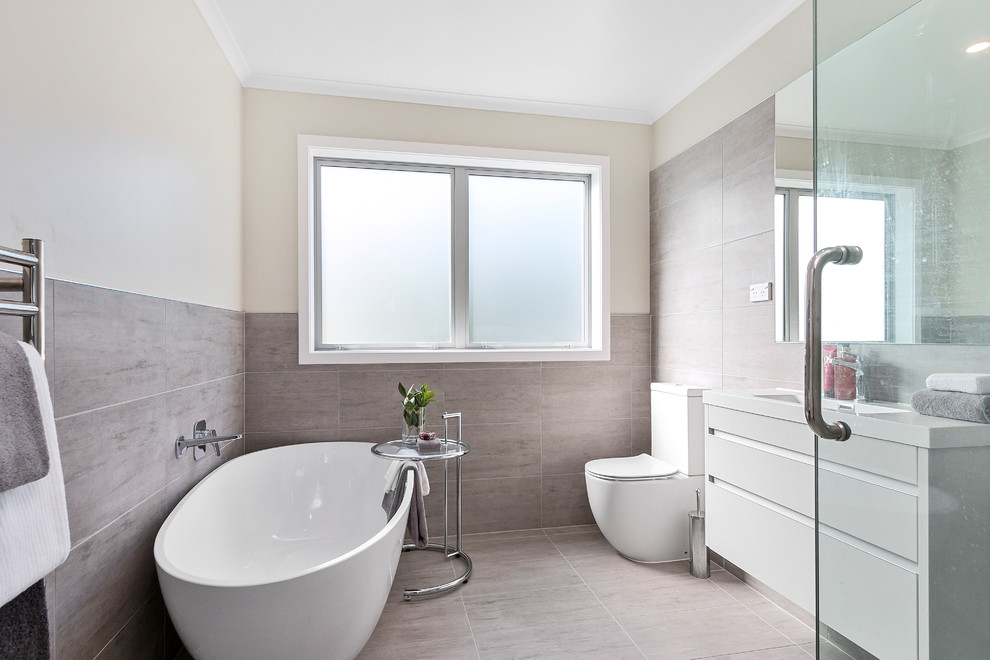 Ejemplo de cuarto de baño actual con puertas de armario blancas, bañera exenta, sanitario de una pieza, baldosas y/o azulejos grises, paredes beige, lavabo suspendido y suelo gris