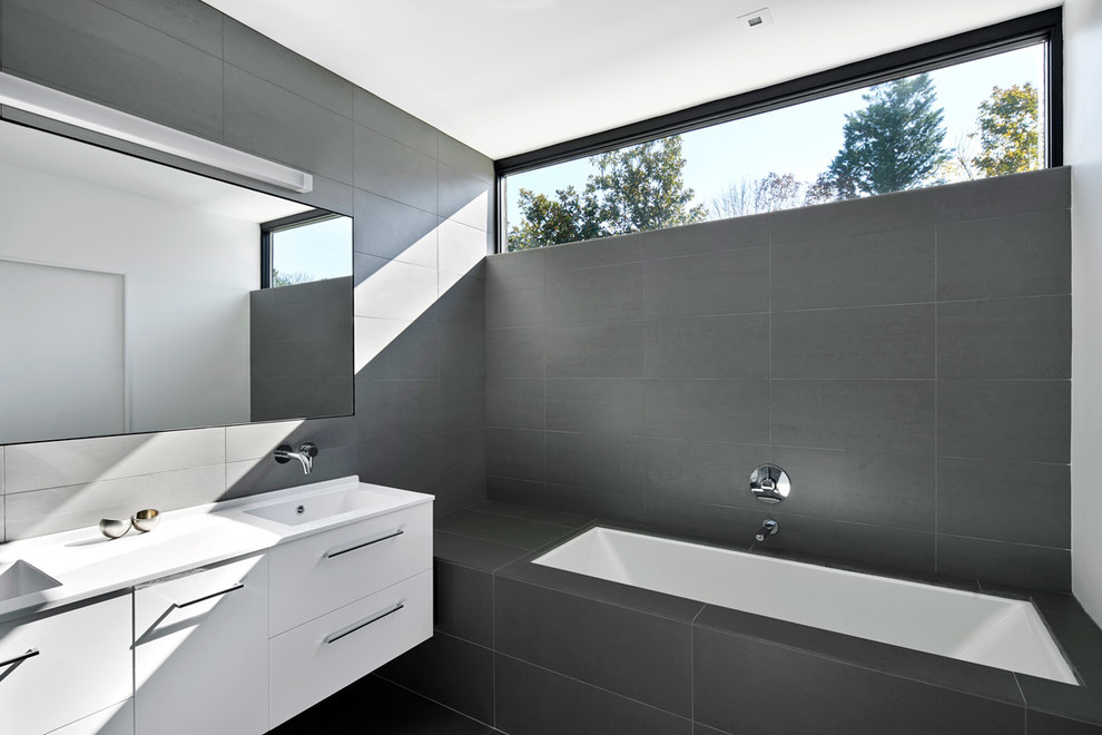 Идея дизайна: главная ванная комната в современном стиле с плоскими фасадами, белыми фасадами, полновстраиваемой ванной, серой плиткой, серыми стенами, монолитной раковиной и белой столешницей