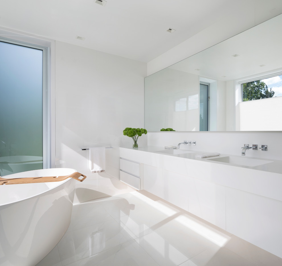 Inredning av ett modernt stort badrum, med släta luckor, vita skåp, ett fristående badkar, vit kakel, vita väggar, klinkergolv i keramik, ett integrerad handfat och bänkskiva i akrylsten