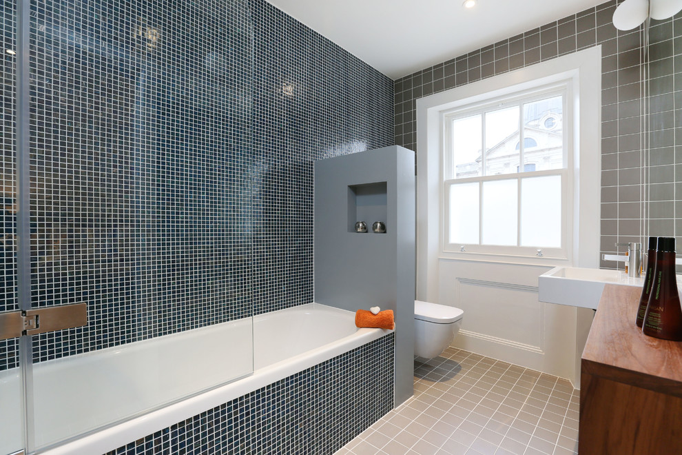 ロンドンにある高級なコンテンポラリースタイルのおしゃれなマスターバスルーム (モザイクタイル、壁付け型シンク、中間色木目調キャビネット、シャワー付き浴槽	、壁掛け式トイレ、黒いタイル、アルコーブ型浴槽) の写真