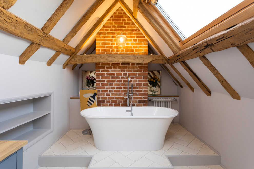 Modelo de cuarto de baño abovedado de estilo de casa de campo con bañera exenta, paredes grises, suelo beige, vigas vistas y ladrillo