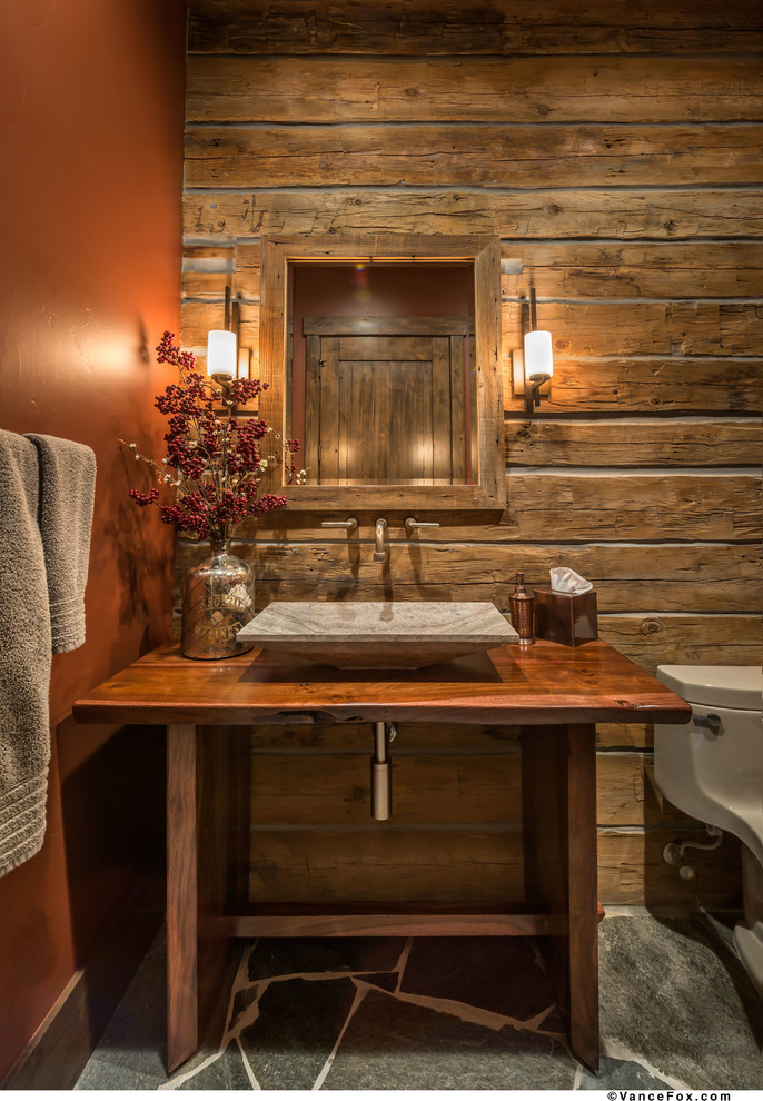 Aménagement d'une grande salle d'eau montagne en bois brun avec une vasque, un placard sans porte, un plan de toilette en bois, WC à poser, un mur orange et un sol en ardoise.