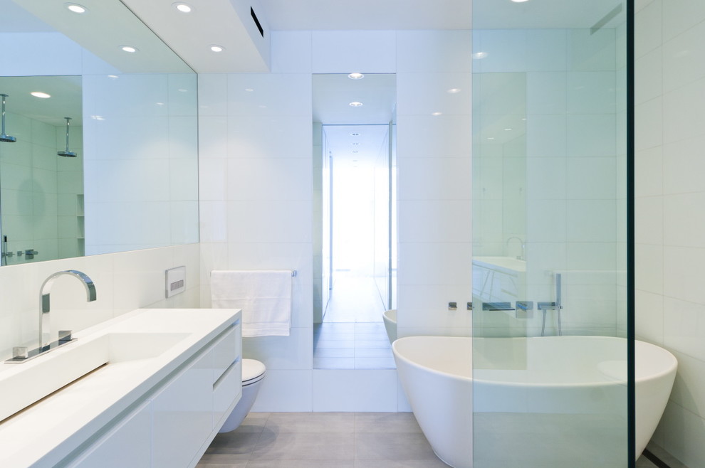 This is an example of a scandi bathroom in New York with a trough sink, flat-panel cabinets, white cabinets, solid surface worktops, a freestanding bath, a built-in shower, a wall mounted toilet, white tiles, white walls and limestone flooring.
