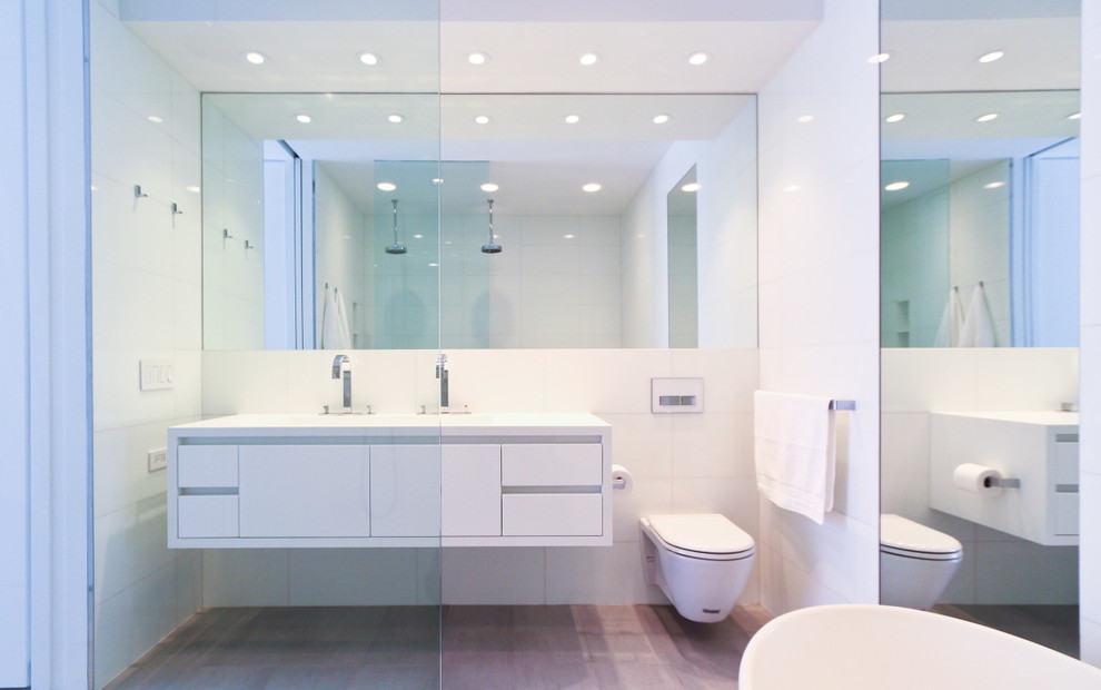 ニューヨークにある北欧スタイルのおしゃれな浴室 (横長型シンク、フラットパネル扉のキャビネット、白いキャビネット、人工大理石カウンター、置き型浴槽、バリアフリー、壁掛け式トイレ、白いタイル、白い壁、ライムストーンの床) の写真