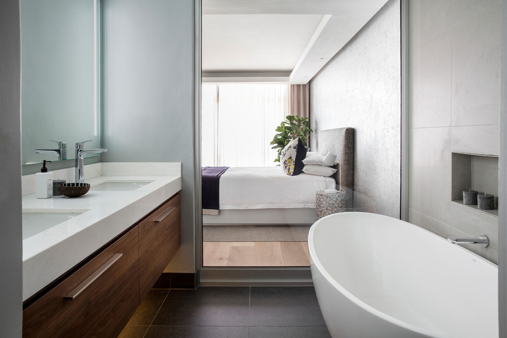 Example of a trendy master gray tile black floor freestanding bathtub design in Other with flat-panel cabinets, dark wood cabinets, gray walls, an undermount sink and white countertops