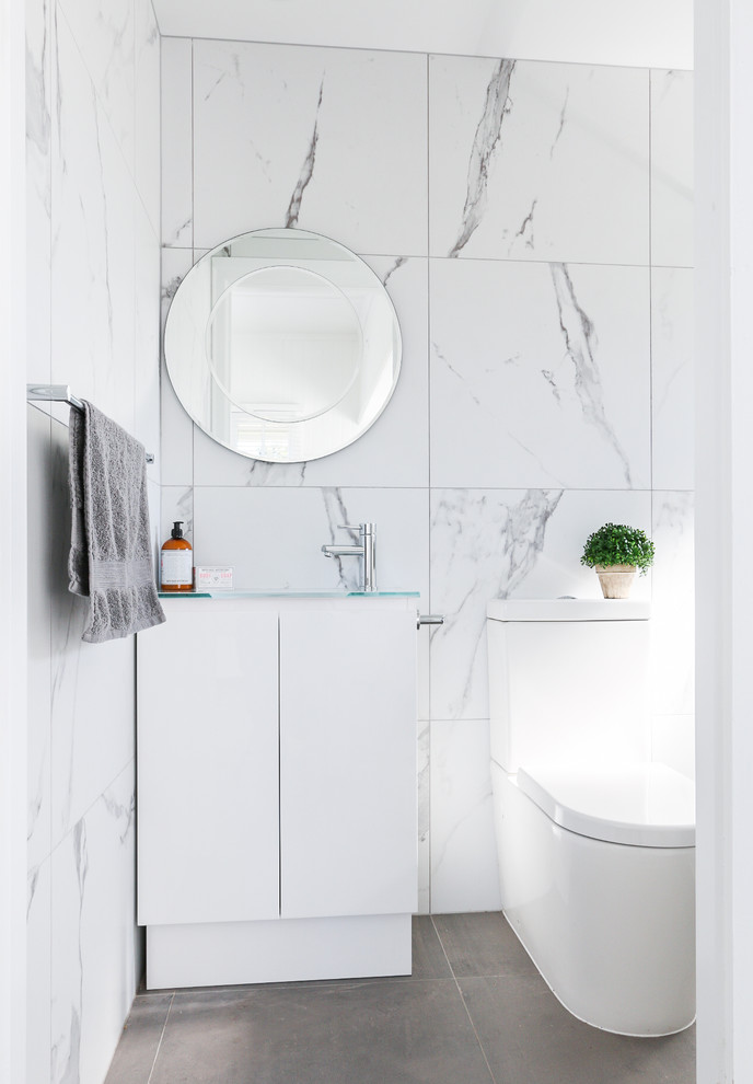 Foto på ett funkis badrum med dusch, med släta luckor, vita skåp, en toalettstol med separat cisternkåpa, vit kakel, marmorkakel, vita väggar, bänkskiva i glas och grått golv