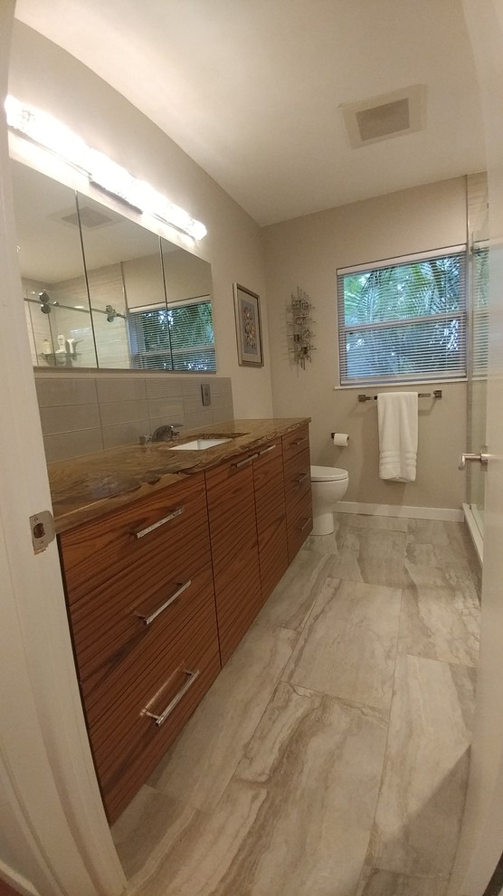 Inspiration for a medium sized contemporary bathroom in Tampa with flat-panel cabinets, medium wood cabinets, an alcove shower, a one-piece toilet, grey tiles, porcelain tiles, grey walls, porcelain flooring, a submerged sink, granite worktops, grey floors, a sliding door and brown worktops.