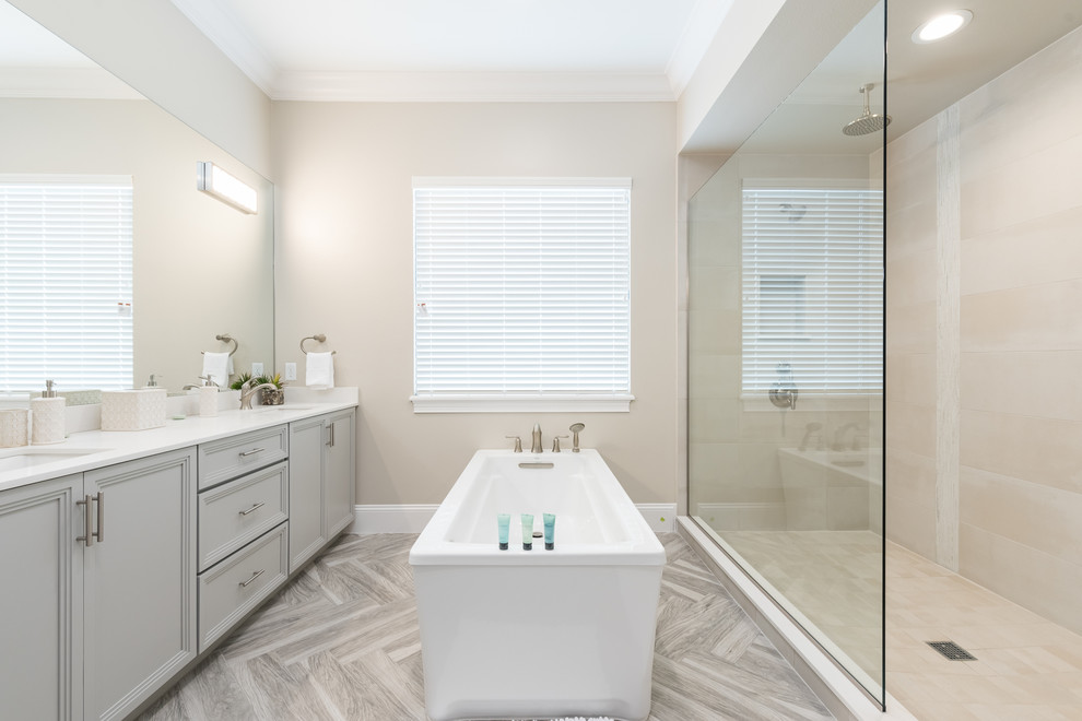 Klassisk inredning av ett badrum, med luckor med infälld panel, grå skåp, ett fristående badkar, en öppen dusch, beige kakel, beige väggar, ett undermonterad handfat, grått golv och med dusch som är öppen
