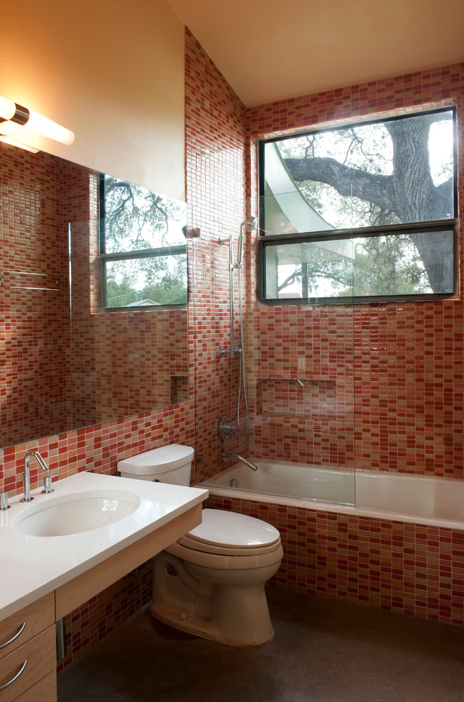 Cette photo montre une petite salle de bain tendance en bois clair avec un lavabo encastré, un placard à porte plane, un plan de toilette en quartz modifié, une baignoire en alcôve, un combiné douche/baignoire, WC à poser, un carrelage rose, un carrelage en pâte de verre, un mur orange et sol en béton ciré.