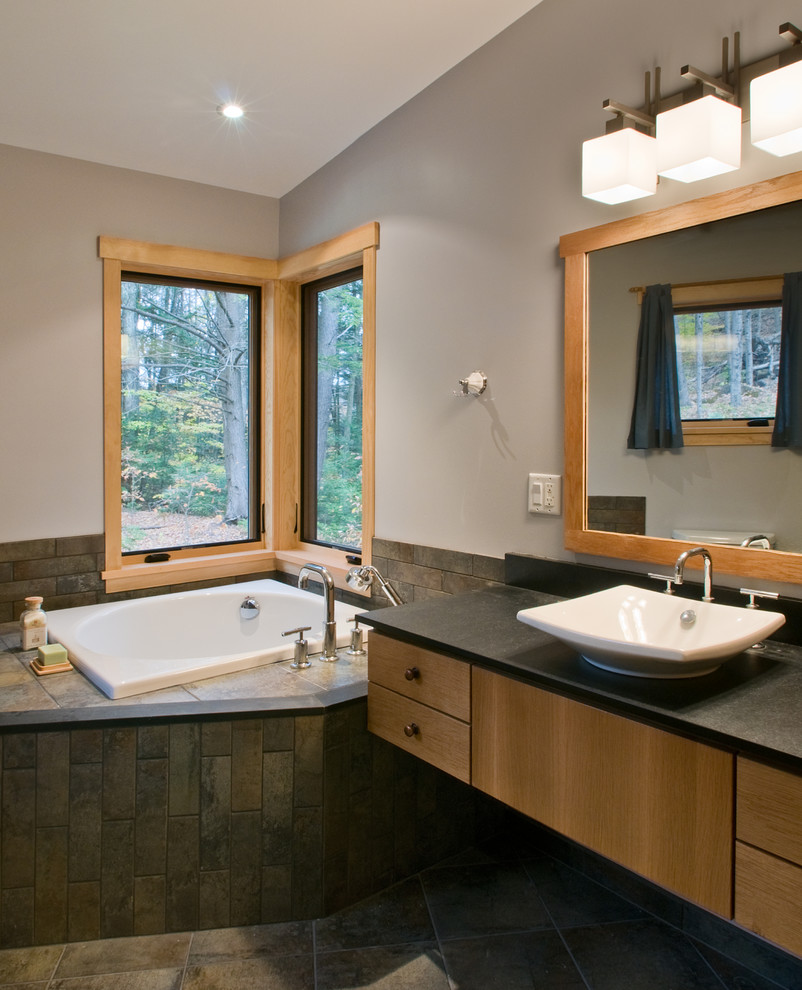 Идея дизайна: большая главная ванная комната в современном стиле с настольной раковиной, светлыми деревянными фасадами, столешницей из оникса, угловой ванной, серыми стенами и полом из керамической плитки