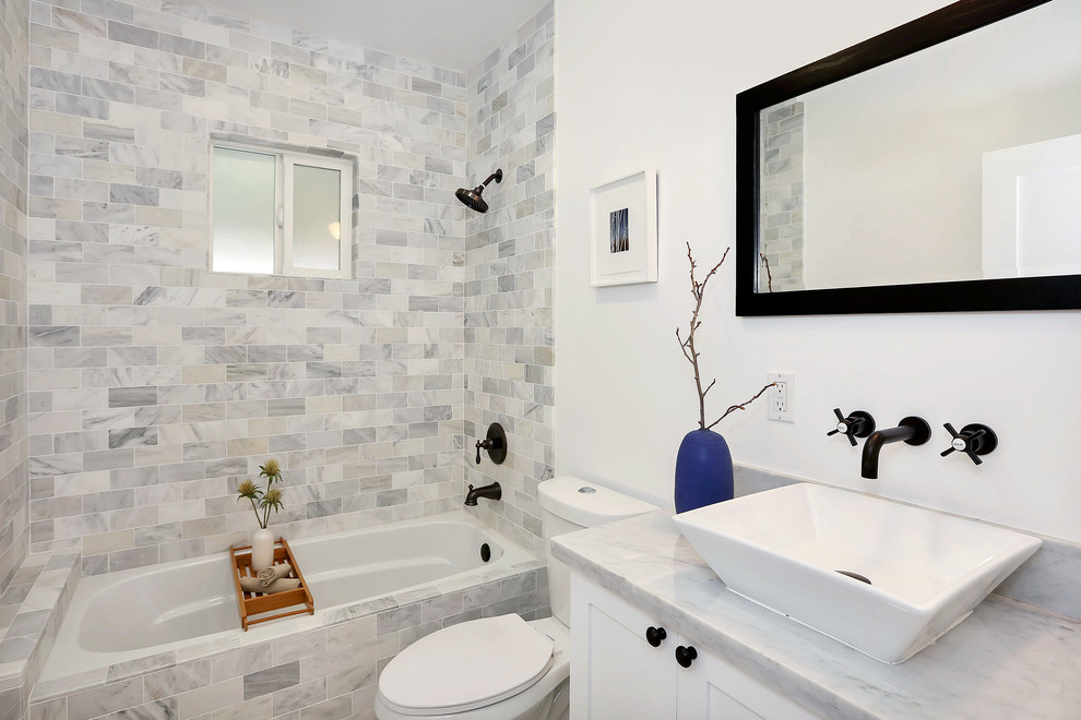 Modelo de cuarto de baño clásico renovado con lavabo sobreencimera, armarios estilo shaker, puertas de armario blancas, bañera empotrada, combinación de ducha y bañera, sanitario de dos piezas y baldosas y/o azulejos grises