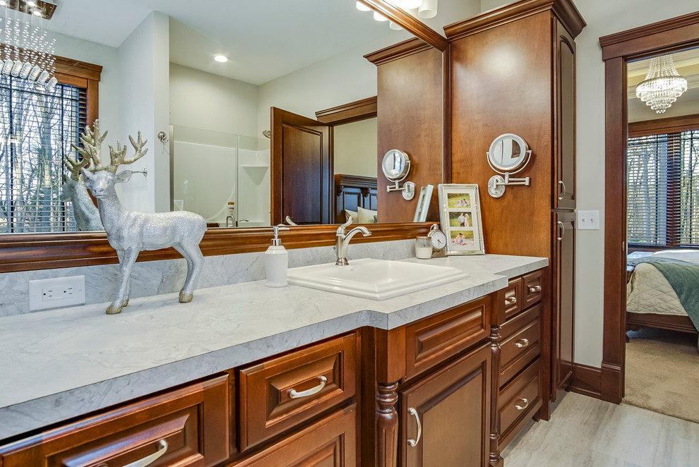 Large elegant master beige floor bathroom photo in Grand Rapids with raised-panel cabinets, brown cabinets, white walls, a drop-in sink, marble countertops, a hinged shower door and white countertops