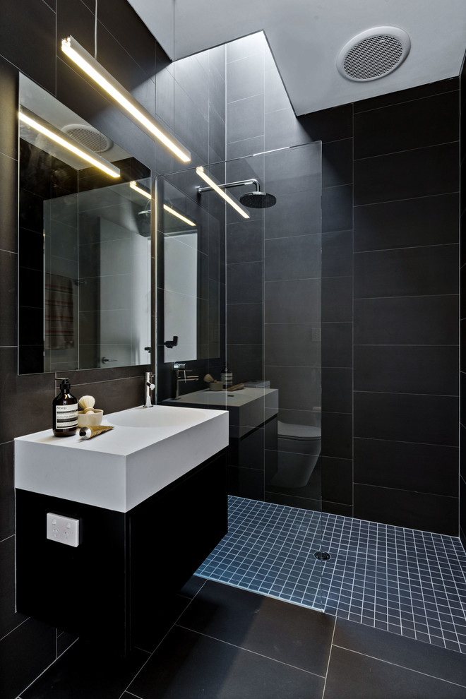 Cette image montre une salle d'eau design avec un placard à porte plane, des portes de placard noires, une douche ouverte, un carrelage noir, un mur noir, un lavabo intégré, un sol noir, aucune cabine et un plan de toilette blanc.