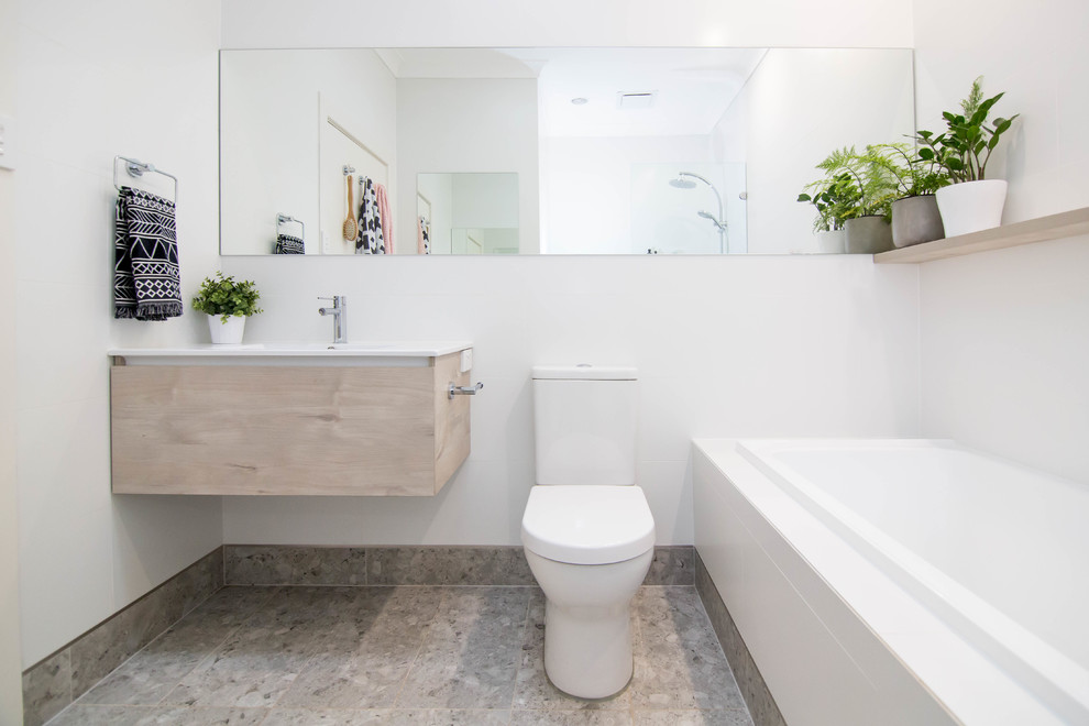 Exemple d'une salle de bain tendance en bois clair avec un placard à porte plane, une baignoire posée, un mur blanc, un plan vasque, un sol gris et un plan de toilette blanc.