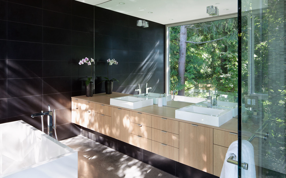 Diseño de cuarto de baño moderno con lavabo sobreencimera, armarios con paneles lisos, puertas de armario de madera clara, encimera de madera, bañera exenta y baldosas y/o azulejos negros
