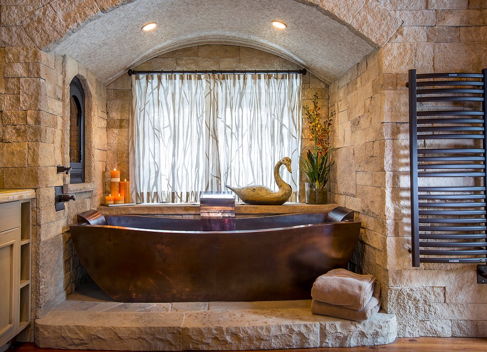 На фото: главная ванная комната в стиле рустика с каменной плиткой и отдельно стоящей ванной с