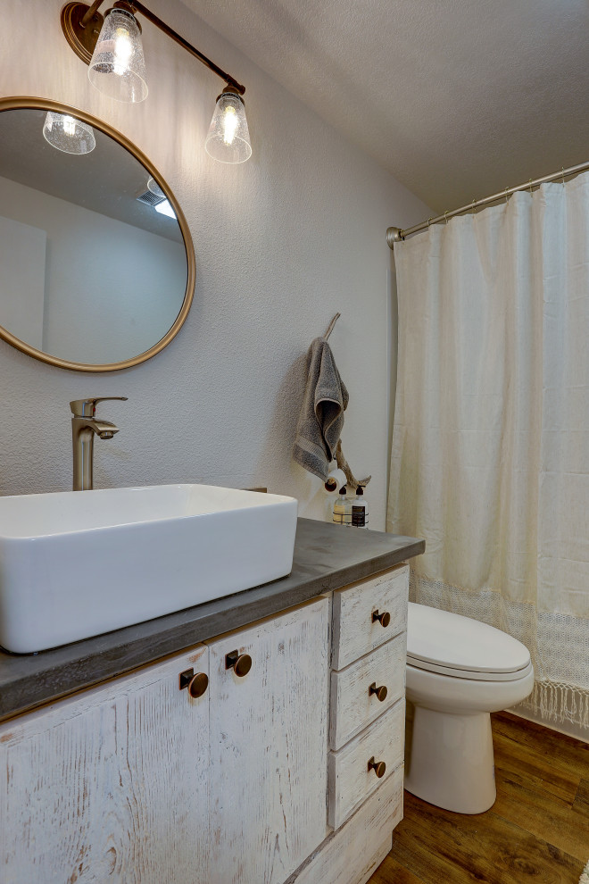 Inredning av ett rustikt litet grå grått badrum, med släta luckor, skåp i slitet trä, ett badkar i en alkov, en toalettstol med hel cisternkåpa, vita väggar, laminatgolv, ett fristående handfat, bänkskiva i akrylsten, brunt golv och dusch med duschdraperi