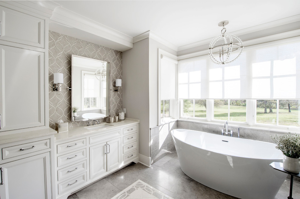Photo of a large traditional ensuite bathroom in Columbus with beaded cabinets, white cabinets, a freestanding bath, grey walls, porcelain flooring, a submerged sink, marble worktops, grey floors and white worktops.