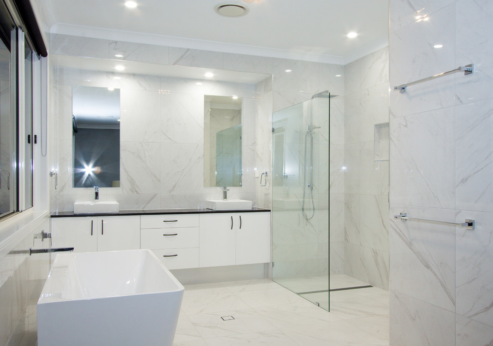 Пример оригинального дизайна: большая главная ванная комната в стиле модернизм с белыми фасадами, отдельно стоящей ванной, открытым душем, белой плиткой, керамогранитной плиткой, белыми стенами, полом из керамогранита, столешницей из гранита, белым полом, открытым душем и черной столешницей