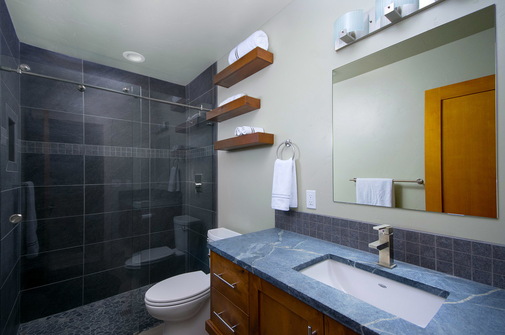 Foto på ett litet industriellt blå badrum, med skåp i shakerstil, skåp i mellenmörkt trä, grå kakel, porslinskakel, grå väggar, klinkergolv i småsten, ett undermonterad handfat, bänkskiva i täljsten, en dusch i en alkov och en toalettstol med separat cisternkåpa