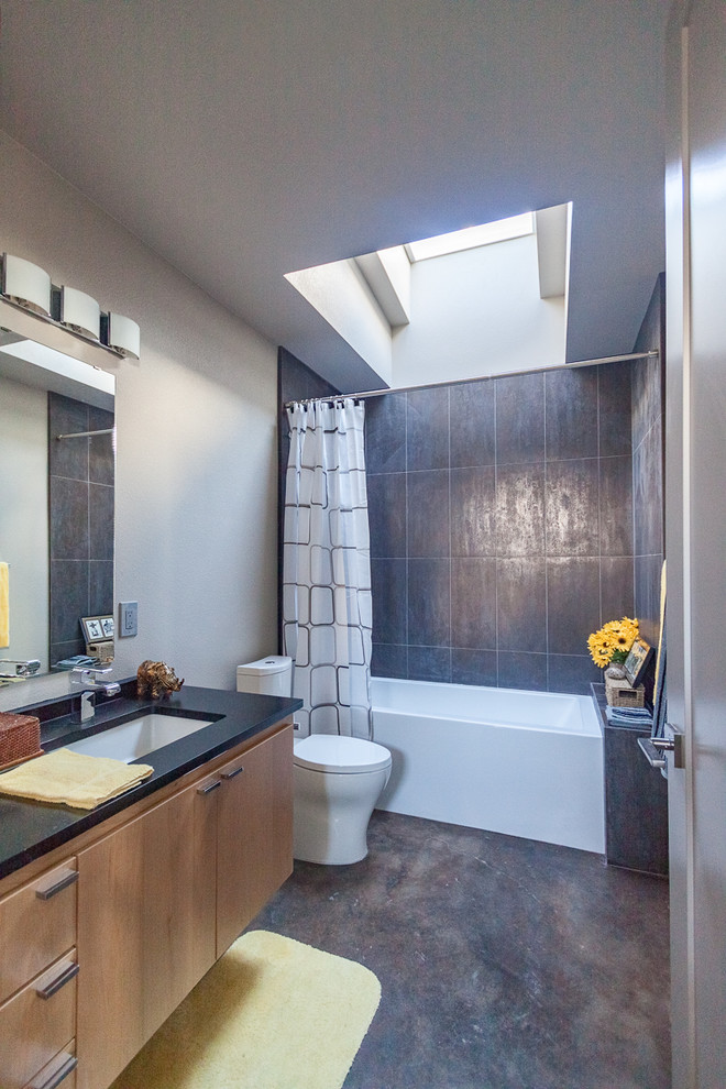 Medium sized contemporary family bathroom in Austin with a submerged sink, flat-panel cabinets, light wood cabinets, granite worktops, an alcove bath, a shower/bath combination, a two-piece toilet, grey tiles, ceramic tiles, beige walls and concrete flooring.