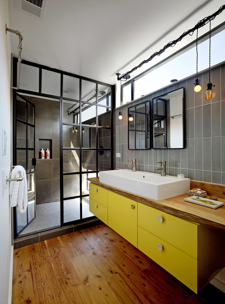 Свежая идея для дизайна: ванная комната среднего размера в стиле лофт с столешницей из дерева, плоскими фасадами, желтыми фасадами, душем в нише, серой плиткой, керамогранитной плиткой, белыми стенами, паркетным полом среднего тона, душевой кабиной, коричневым полом, душем с распашными дверями и коричневой столешницей - отличное фото интерьера