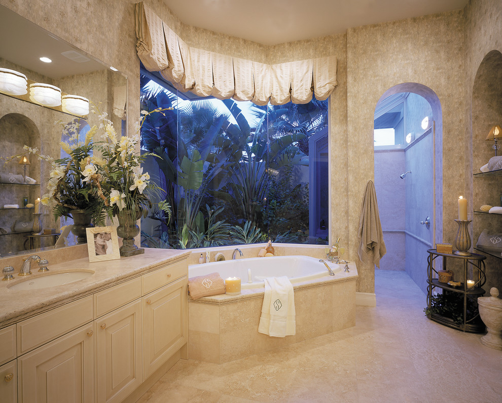 Photo of a large mediterranean ensuite bathroom in Miami with a submerged sink, raised-panel cabinets, light wood cabinets, marble worktops, a built-in bath, a walk-in shower, a two-piece toilet, beige tiles, stone tiles, beige walls and travertine flooring.