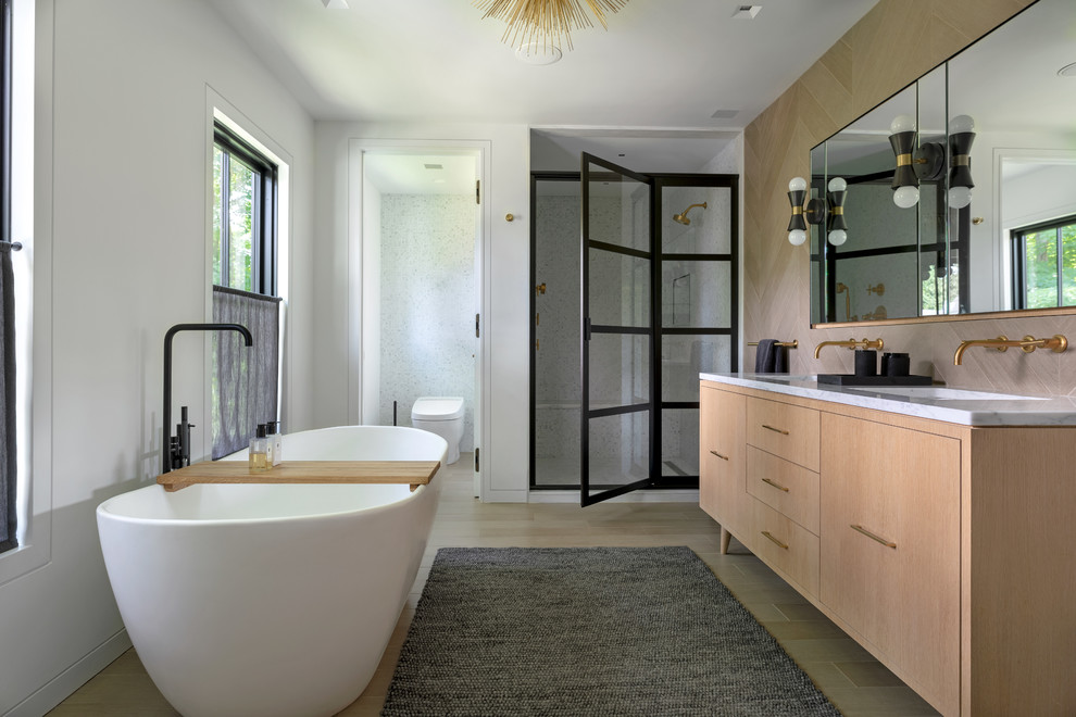 Photo of a rural ensuite bathroom in New York with light wood cabinets, a freestanding bath, an alcove shower, white walls, a submerged sink, beige floors, a hinged door, grey worktops and flat-panel cabinets.