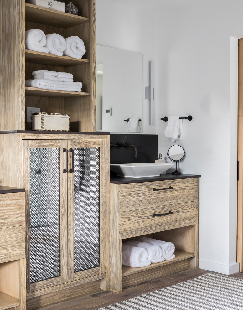 Diseño de cuarto de baño rural con puertas de armario de madera clara, paredes blancas, suelo de madera oscura, lavabo sobreencimera y encimeras negras