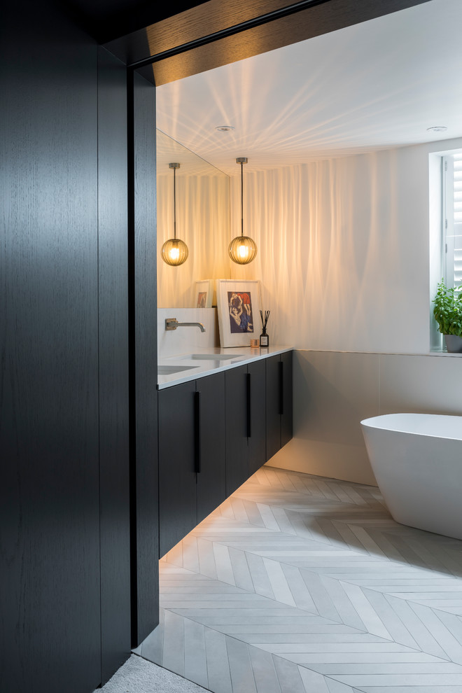 Modernes Badezimmer mit grauem Boden, flächenbündigen Schrankfronten, schwarzen Schränken, freistehender Badewanne, beigen Fliesen, weißer Wandfarbe, integriertem Waschbecken und weißer Waschtischplatte in London