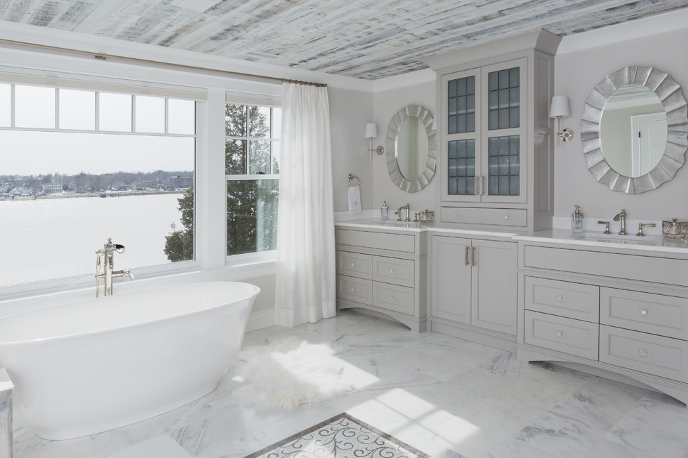 Foto de cuarto de baño costero con armarios con rebordes decorativos, puertas de armario grises, bañera exenta, paredes grises, lavabo bajoencimera, suelo gris y encimeras blancas
