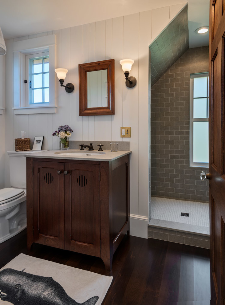 Bild på ett litet lantligt badrum med dusch, med luckor med infälld panel, skåp i mörkt trä, en öppen dusch, grå kakel, cementkakel, vita väggar, mörkt trägolv, bänkskiva i kalksten, ett undermonterad handfat och en toalettstol med hel cisternkåpa