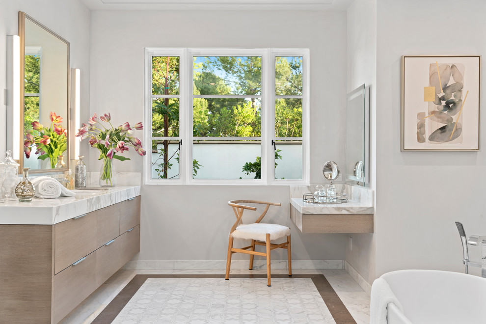 Diseño de cuarto de baño actual con armarios con paneles lisos, puertas de armario de madera clara, bañera exenta, paredes grises, lavabo bajoencimera, suelo blanco, encimeras blancas y espejo con luz