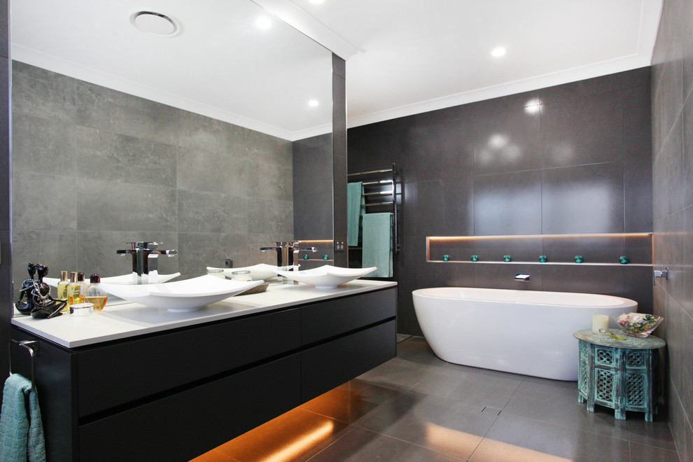 На фото: главная ванная комната среднего размера в стиле модернизм с серыми фасадами, отдельно стоящей ванной, инсталляцией, серой плиткой, керамогранитной плиткой, серыми стенами, полом из керамогранита, настольной раковиной и столешницей из искусственного кварца
