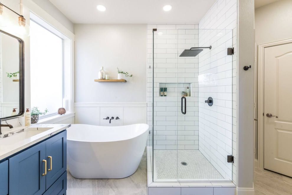 Идея дизайна: главная ванная комната среднего размера в стиле неоклассика (современная классика) с фасадами в стиле шейкер, синими фасадами, отдельно стоящей ванной, угловым душем, белой плиткой, керамической плиткой, серыми стенами, полом из керамогранита, врезной раковиной, столешницей из искусственного кварца, серым полом, душем с распашными дверями, белой столешницей, нишей, тумбой под две раковины, встроенной тумбой и панелями на части стены