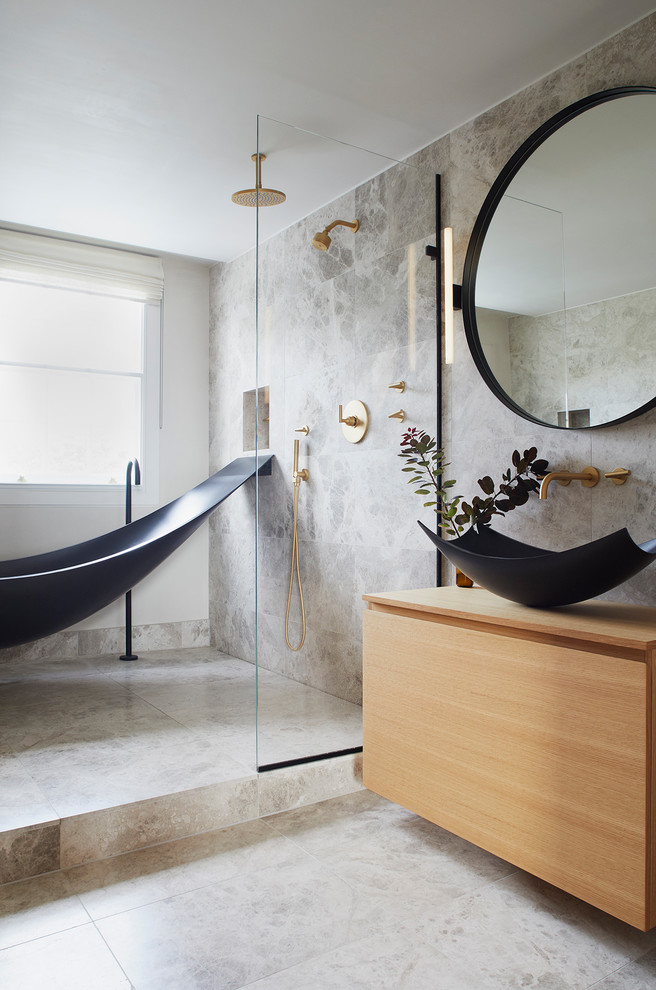 This is an example of a medium sized contemporary ensuite wet room bathroom in London with light wood cabinets, a freestanding bath, a wall mounted toilet, grey tiles, limestone tiles, grey walls, limestone flooring, a vessel sink, wooden worktops, grey floors, an open shower, beige worktops and flat-panel cabinets.