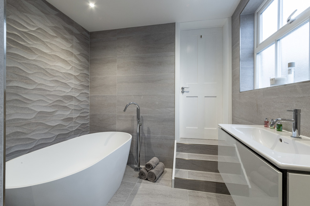 Medium sized contemporary ensuite bathroom in Surrey with louvered cabinets, white cabinets, a freestanding bath, a corner shower, a wall mounted toilet, grey tiles, porcelain tiles, grey walls, porcelain flooring, a wall-mounted sink, solid surface worktops, grey floors, a hinged door and white worktops.