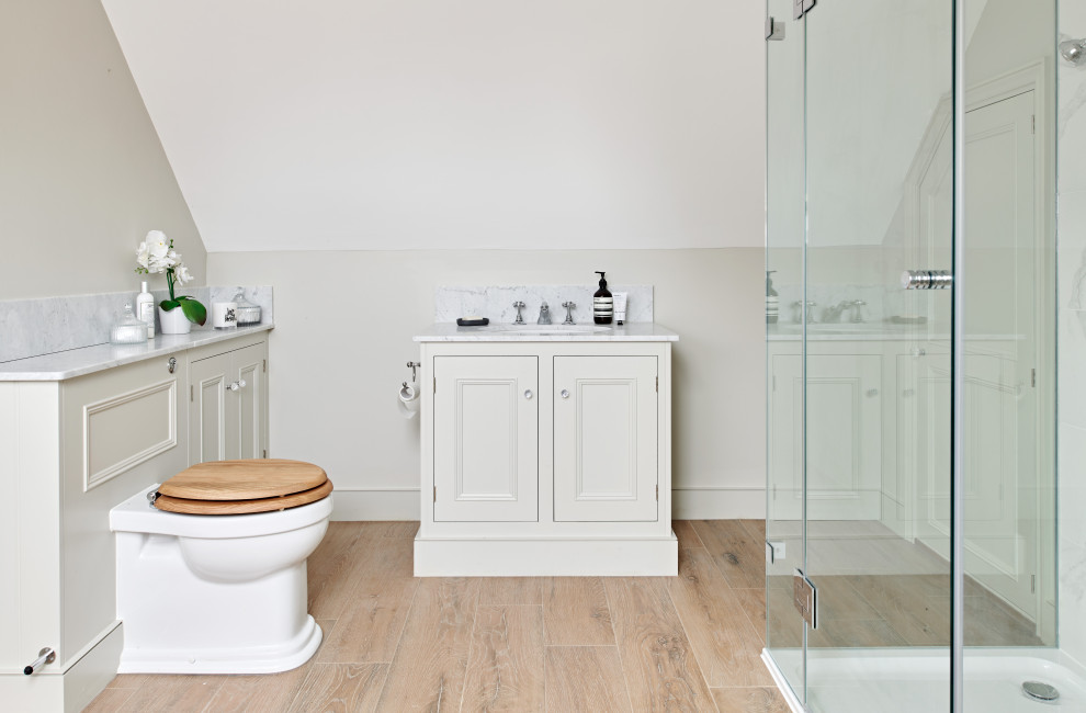 Klassisches Badezimmer mit Schrankfronten mit vertiefter Füllung, beigen Schränken, Eckdusche, beiger Wandfarbe, braunem Holzboden, Unterbauwaschbecken, braunem Boden, Falttür-Duschabtrennung und weißer Waschtischplatte in Wiltshire
