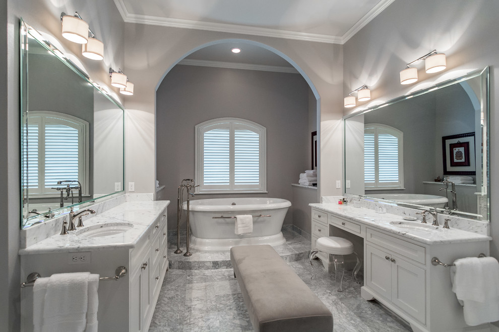 На фото: большая главная ванная комната в классическом стиле с фасадами с утопленной филенкой, белыми фасадами, отдельно стоящей ванной, мраморным полом, врезной раковиной и мраморной столешницей
