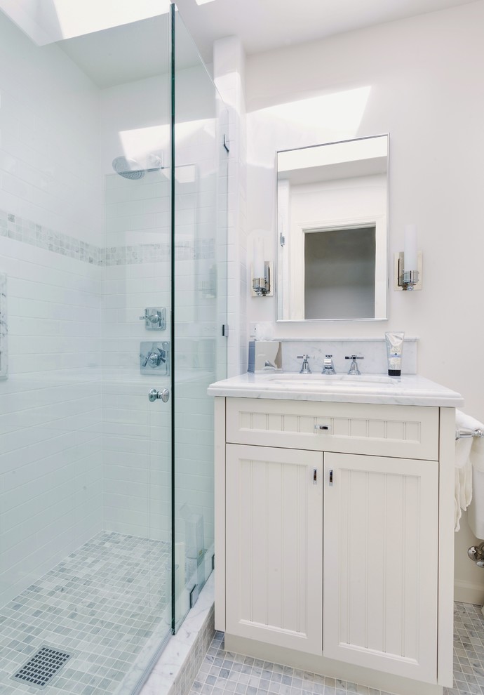 Maritim inredning av ett litet badrum med dusch, med ett undermonterad handfat, luckor med profilerade fronter, vita skåp, marmorbänkskiva, vit kakel, stenkakel, vita väggar och mosaikgolv