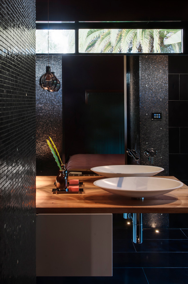 Photo of a small modern ensuite bathroom in Sydney with a vessel sink, flat-panel cabinets, white cabinets, wooden worktops, a corner shower, a wall mounted toilet, black tiles, mosaic tiles, black walls and ceramic flooring.