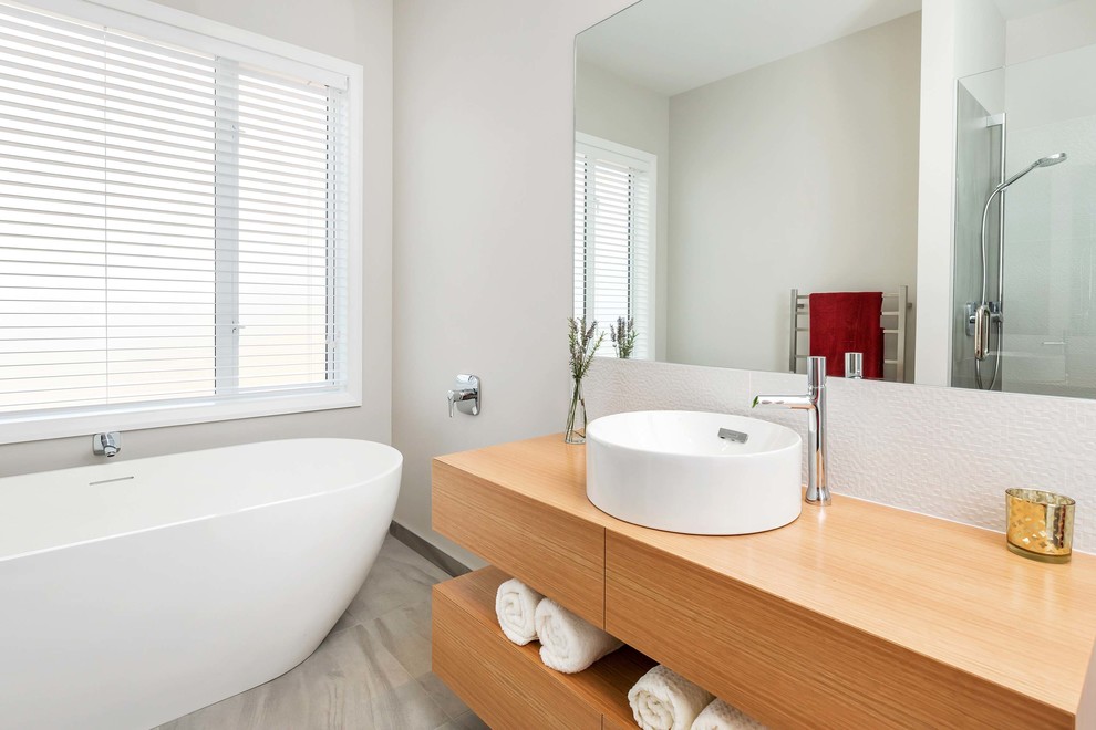 Exemple d'une grande salle de bain tendance en bois brun avec une baignoire indépendante, un carrelage beige, un carrelage de pierre, un plan de toilette en bois, un placard à porte plane, un mur gris, une vasque et un plan de toilette marron.