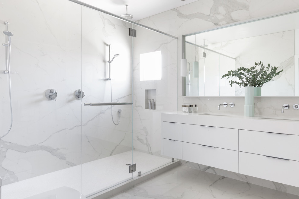 Modern inredning av ett vit vitt badrum, med släta luckor, vita skåp, en dusch i en alkov, vit kakel, stenhäll, ett undermonterad handfat, vitt golv och dusch med gångjärnsdörr