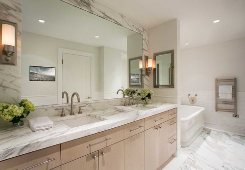 Light wood bathroom cabinets with flat-panel drawers, long pulls, marble countertop, double sinks, large mirror, and soaking tub.