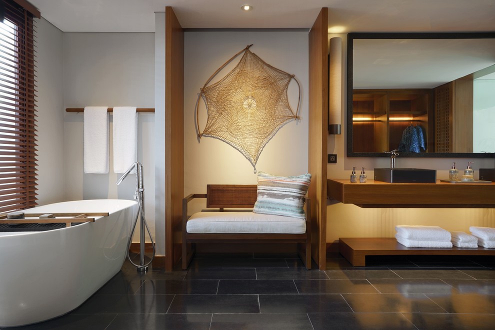 Aménagement d'une salle de bain principale exotique en bois foncé avec un placard sans porte, une baignoire indépendante, un mur blanc, une vasque, un plan de toilette en bois et un sol noir.