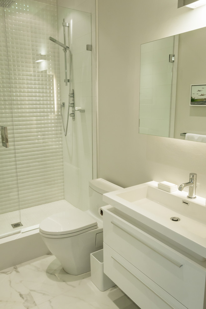 Bild på ett mellanstort funkis badrum med dusch, med ett integrerad handfat, släta luckor, vita skåp, bänkskiva i akrylsten, en toalettstol med hel cisternkåpa, vit kakel, porslinskakel, en dusch i en alkov, grå väggar och marmorgolv