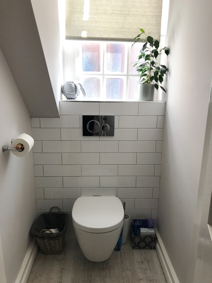 Bild på ett litet maritimt toalett, med släta luckor, vita skåp, en vägghängd toalettstol, grå kakel, porslinskakel, grå väggar, vinylgolv och ett väggmonterat handfat