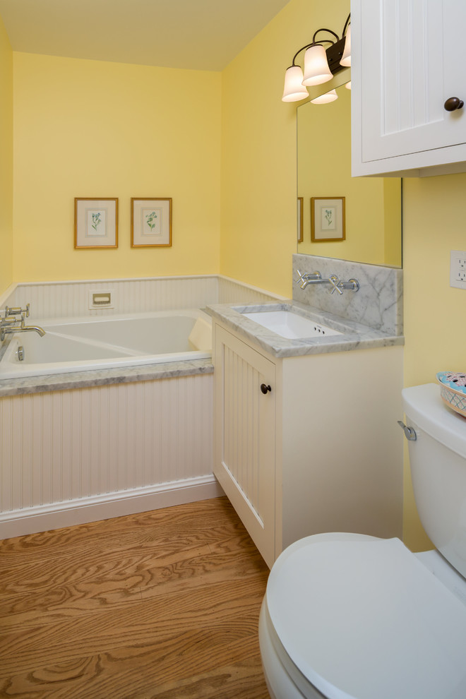 Idées déco pour une petite salle d'eau classique avec un placard à porte shaker, des portes de placard blanches, une baignoire posée, WC séparés, un carrelage blanc, un mur jaune, un sol en bois brun, un lavabo encastré et un plan de toilette en marbre.