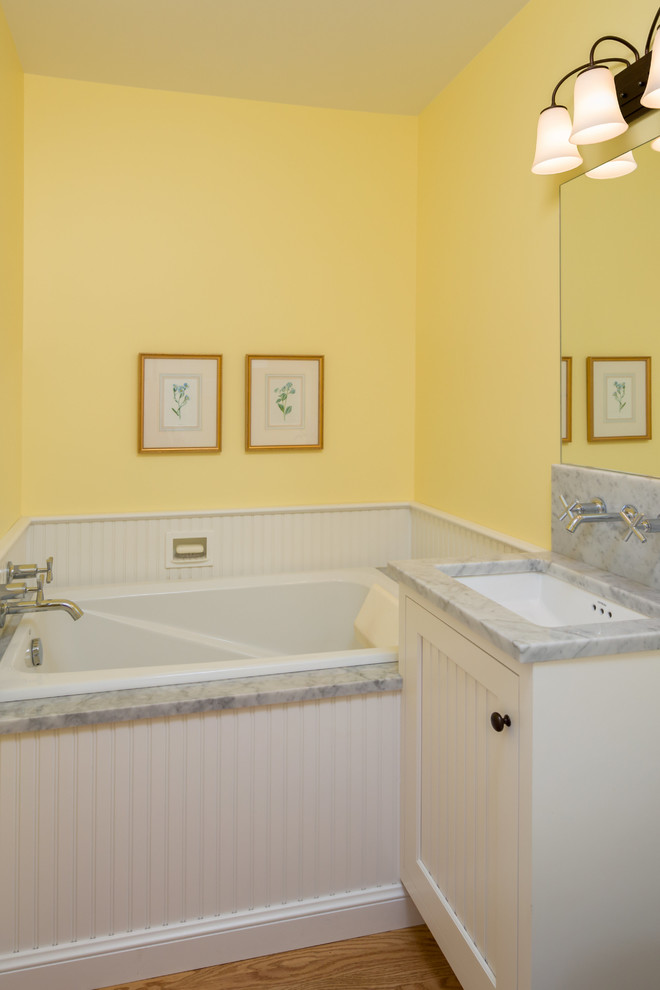 Cette photo montre une petite salle d'eau chic avec des portes de placard blanches, une baignoire posée, WC séparés, un mur jaune, un lavabo encastré, un plan de toilette en marbre, un placard à porte shaker, un carrelage blanc et un sol en bois brun.