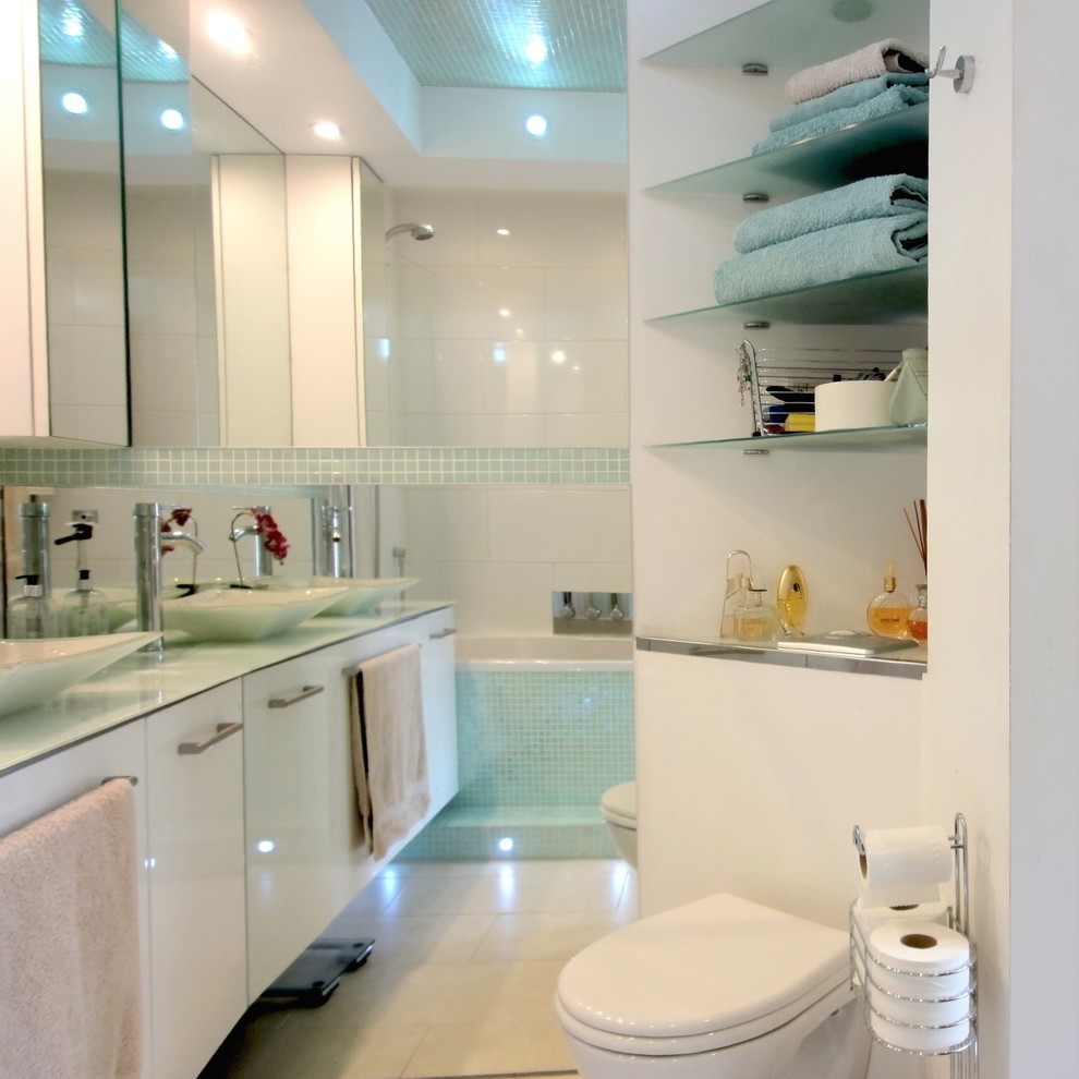 Cette image montre une petite salle de bain minimaliste avec une vasque, un placard à porte plane, des portes de placard blanches, un plan de toilette en verre, une baignoire en alcôve, un carrelage vert, mosaïque, un mur blanc et un sol en carrelage de céramique.